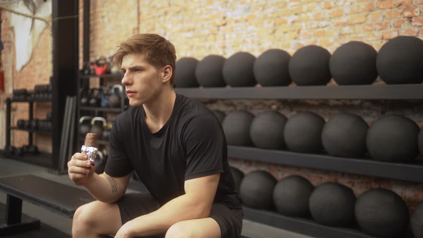 Focused young athlete in black athletic wear consuming protein bar after intense workout in industrial gym