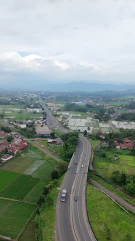 Aerial view of a countryside highway curving through rice fields and small houses, with distant hills and cloudy blue sky creating a peaceful rural landscape