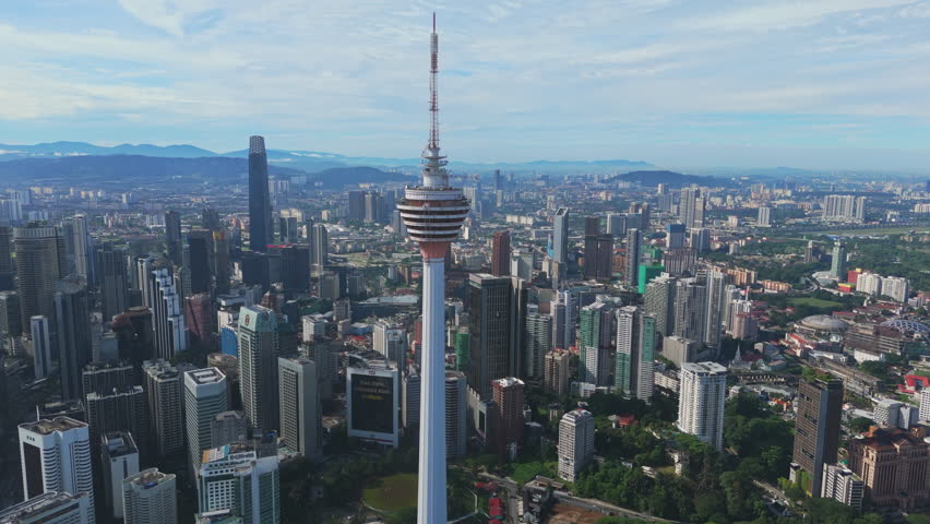 KL Tower dominates Kuala Lumpur, Malaysia as the drone glides above a dense skyline and green parkland in bright daylight. Morning clarity highlights business energy, architecture and tourism appeal