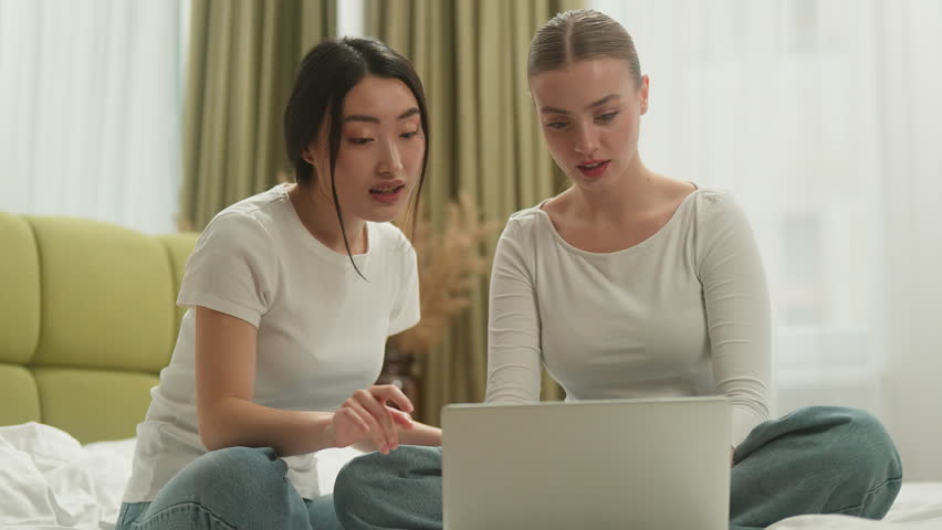 Two multiracial happy women winning with laptop casino bet victory amazed on bed with computer smiling laughing multiethnic friends Caucasian Asian girls rejoice cuddling win high five celebration