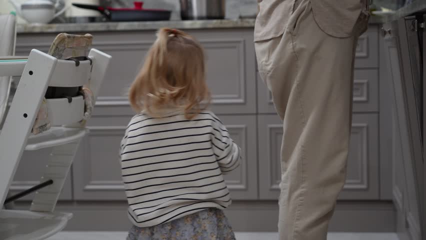 Curious child in striped shirt and ponytail plays with toy phone while observing adult in calm, modern kitchen, capturing innocent, everyday moments of domestic life.
