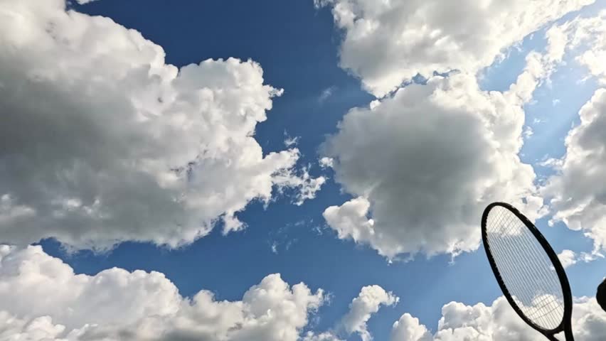 Badminton racket hits shuttlecock against blue sky with bright clouds. Quick upward motion captures flying object and dynamic sunlight reflections.