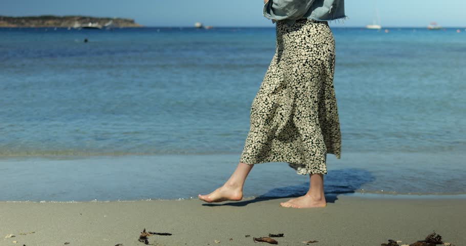 Woman walks along wet sand near sea in patterned skirt and denim jacket. Walker lifts heel and sets toes in shallow water keeping balance as step advances