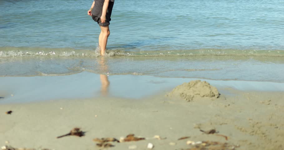 Woman stands shin deep in sea near shore in shorts and t-shirt. Female tourist faces water and lets small wave reach legs waiting near beach wet sand