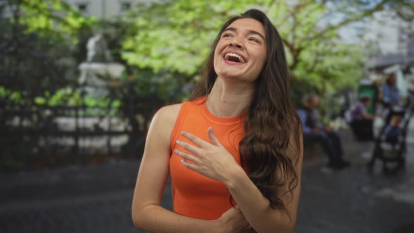 Young hispanic woman wearing orange tank top with hand on chest smiling in forest at midday sun; joy.