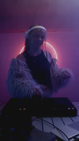 Vertical: Young beauty woman dancing on Dj set in red night techno club. Modern pretty female wear in pink fur coat. Musician artist make trance music at a rave party. Neon blue light, white earphones