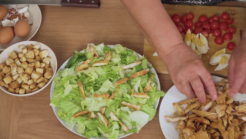 Placing grilled chicken strips onto lettuce base for Caesar salad. Adding cooked poultry pieces on fresh greens for salad preparation. Arranging seasoned chicken over leafy lettuce for Caesar dish