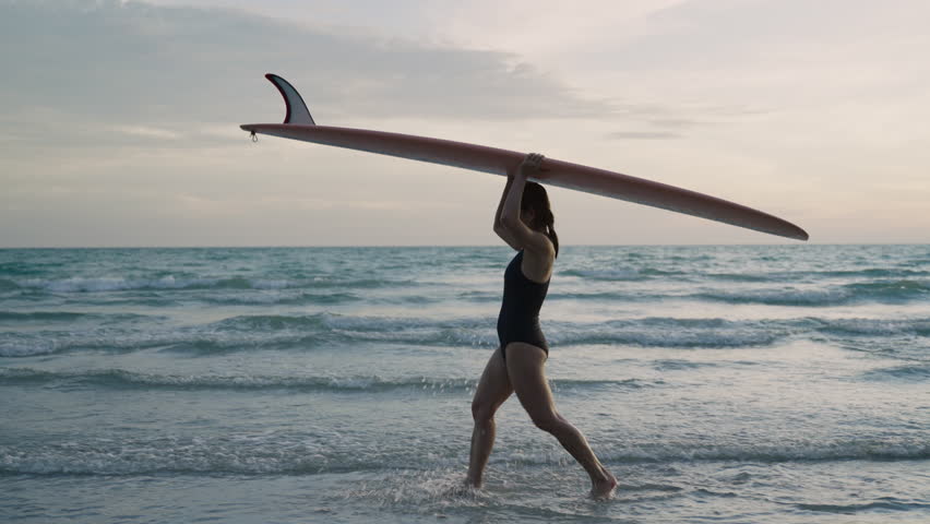 Asian woman surfer in a bikini holding a surfboard on her head and walking with happiness on the beach after playing surfing during summer vacation holiday at the coastline. 