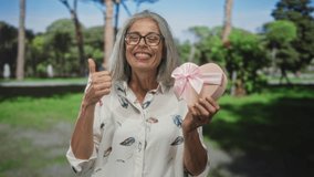 Woman holding heart shaped gift box and gives thumbs up gesture in a leafy park with trees and a walking path; happiness gratitude. - Powered by Shutterstock - Get 15% off with code: PIKWIZARD15