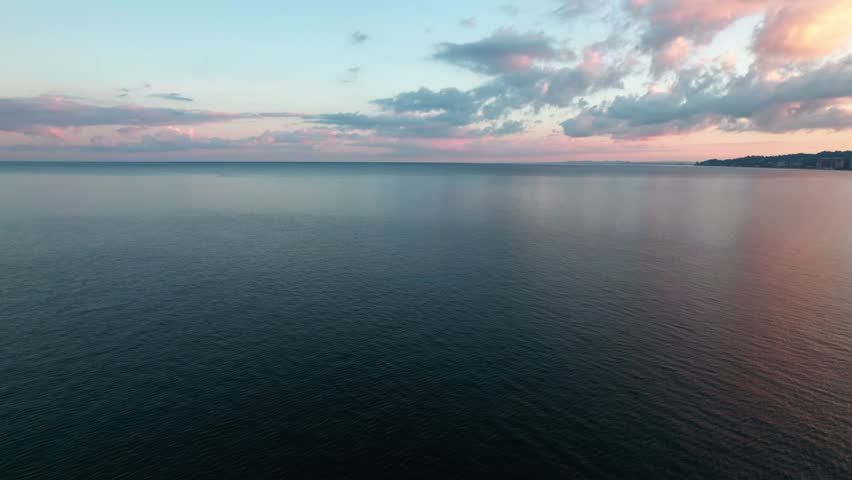 Aerial drone view of the Black Sea at sunrise. Calm water, soft pink clouds on the horizon, and deep morning colors create a peaceful, cinematic seascape