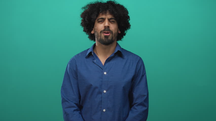 Man with curly hair and clasped hands wearing a blue shirt and wristwatch, hands to chest gesture in studio with teal backdrop; sincere gratitude.