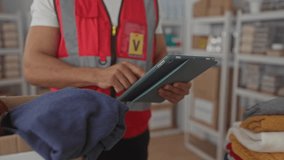Man tapping tablet while holding folded clothes near a donation box, wearing red volunteer vest and badge in a building donation center; compassion charity organizing. - Powered by Shutterstock - Get 15% off with code: PIKWIZARD15