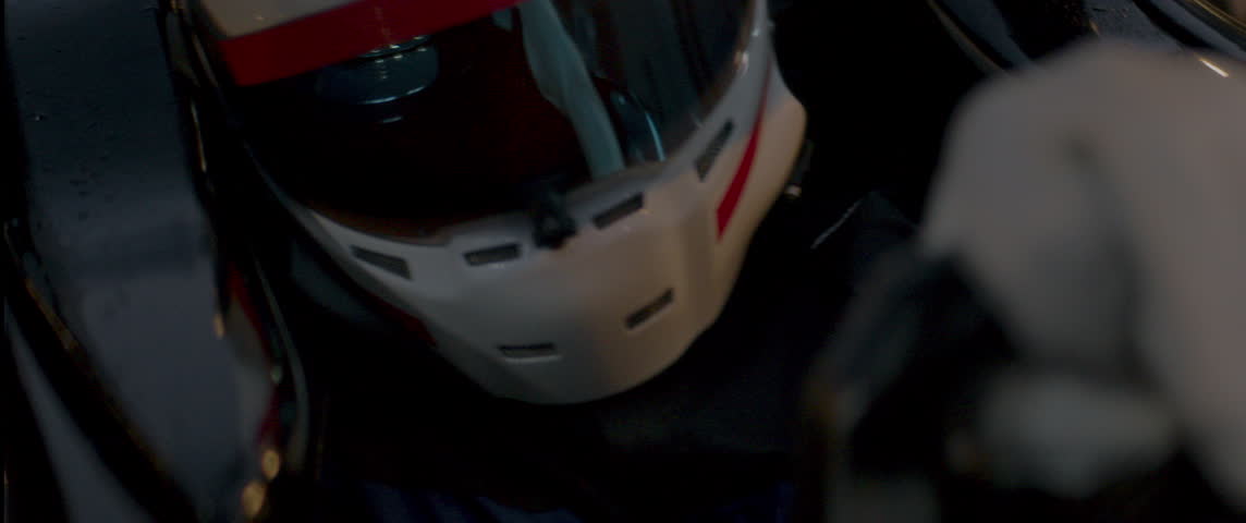Close-up of a race car driver in a white helmet with red details, driving fast on a racing track, evening race