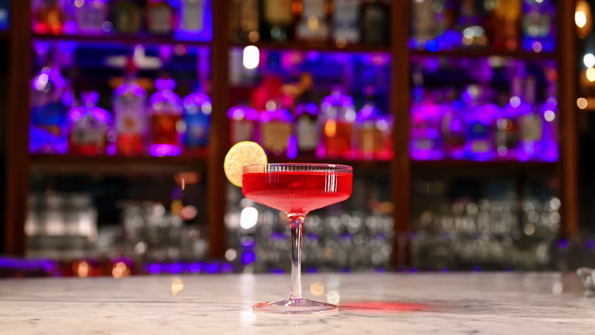 Red cocktail with lime slice on marble bar counter. Elegant red cocktail with lime garnish served in coupe glass at neon-lit bar counter at night.