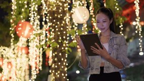 Portrait of focused Asian woman browses social network on digital tablet under tree with fairy lights. Korean lady uses device and smiles on night downtown street slow motion - Powered by Shutterstock - Get 15% off with code: PIKWIZARD15