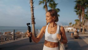 Fitness blogger woman films herself while walking outdoors on a sunny morning. She holds a camera in one hand and carries a yoga mat over her shoulder. Concept of active lifestyle and content creation - Powered by Shutterstock - Get 15% off with code: PIKWIZARD15