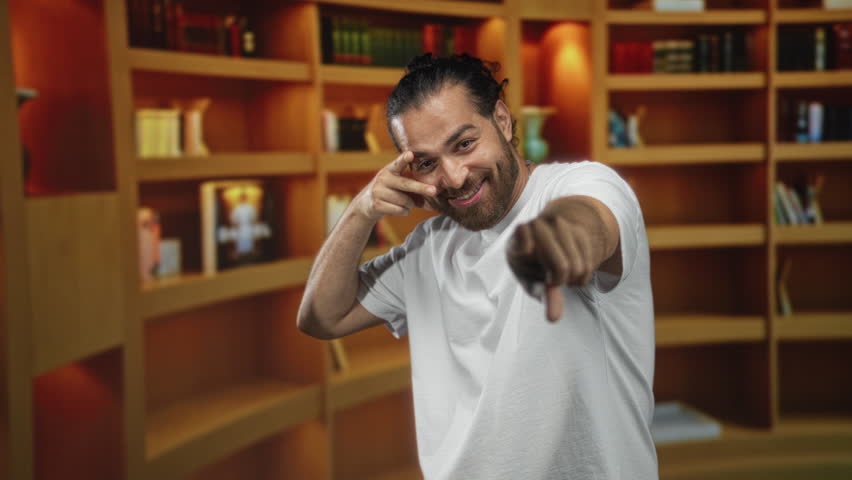 Young arab hispanic man points finger to camera while smiling, wearing white tshirt in library with books and wooden shelves; playful confidence.