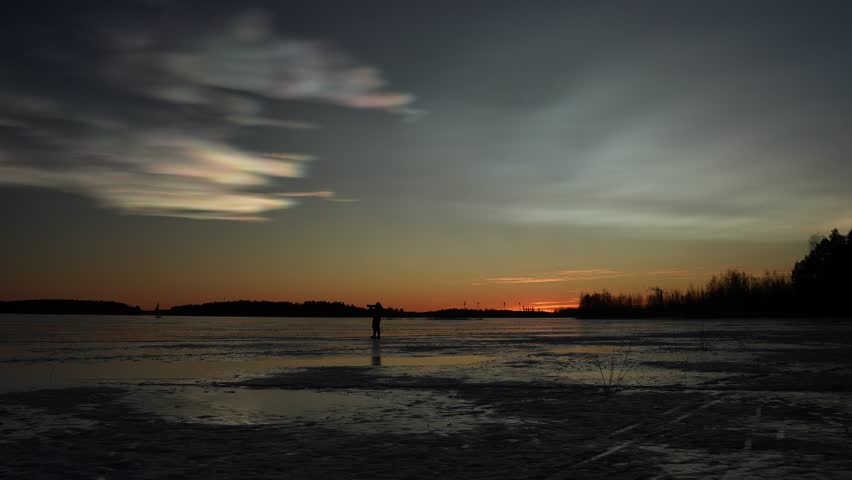 Photographer walking across frozen lake during sunset with colorful clouds and warm evening light in peaceful Finnish winter scenery.