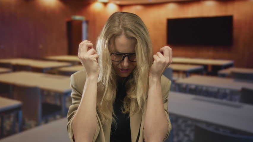 Woman expressing frustration in classroom with blonde hair, glasses, beige blazer, focusing on emotion in educational indoor setting with wood background.
