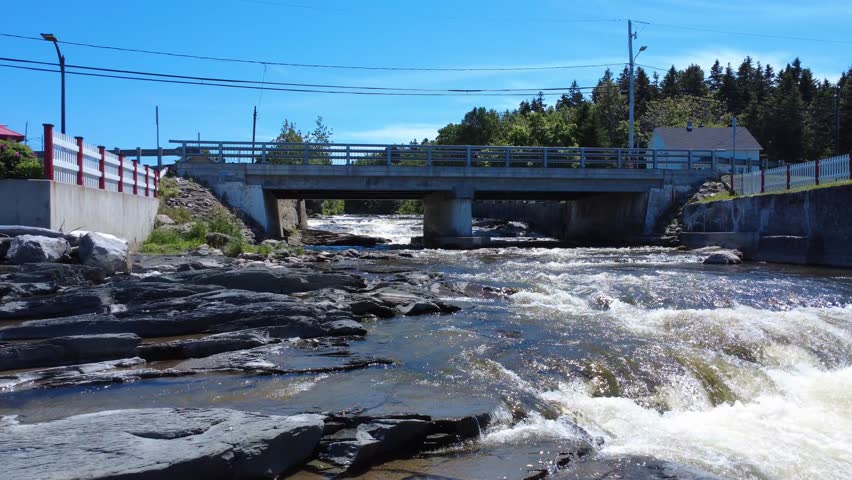Low-level flight over a cascading river passing under a small rural concrete road bridge to its central pillar. Riviere-Blanche, Saint-Ulric-de-Matane, Quebec, Canada, 2025.