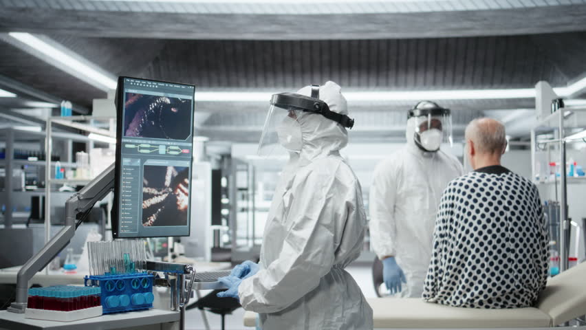 Doctor in quarantine performing clinical study on an infectious patient, testing vaccine responses and monitoring DNA molecules on monitor to develop treatments for epidemics. Camera A.
