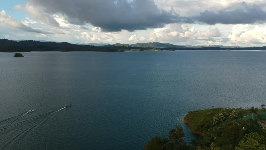 Drone wide of a tropical reservoir with forested islets and floating fish-farm platforms. Soft evening light and sun rays on rippled water; shoreline jetty in foreground. No people or logos; 