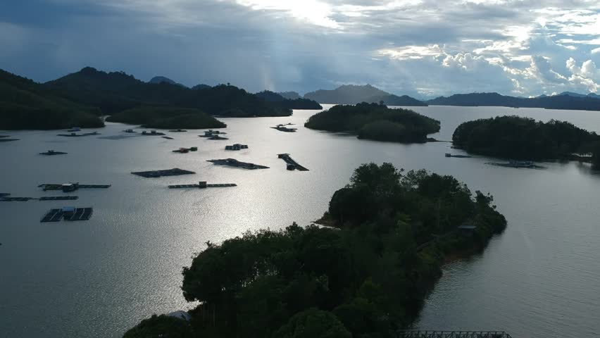 Drone wide of a tropical reservoir with forested islets and floating fish-farm platforms. Soft evening light and sun rays on rippled water; shoreline jetty in foreground. No people or logos; 