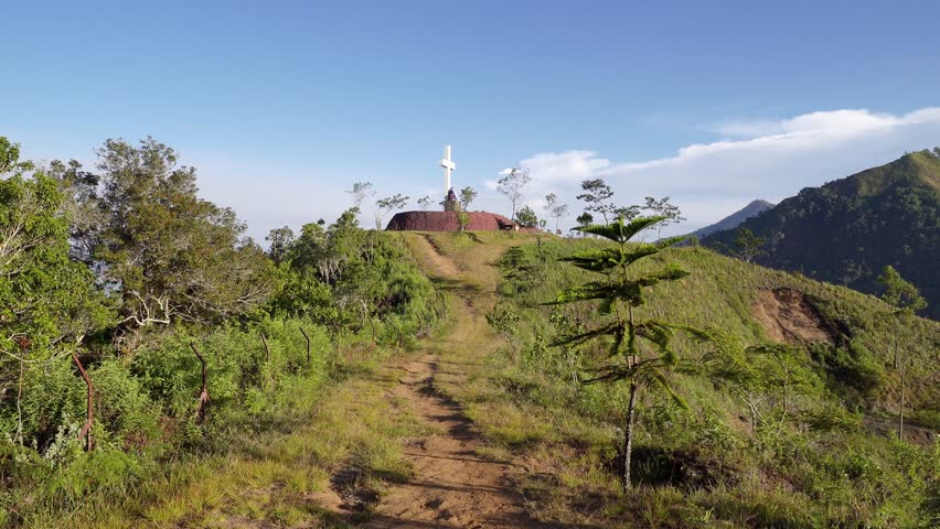 An immersive POV video walking up a dirt path on a lush green hill towards a white Christian cross at the summit. Represents a spiritual journey, pilgrimage, or reaching a destination.