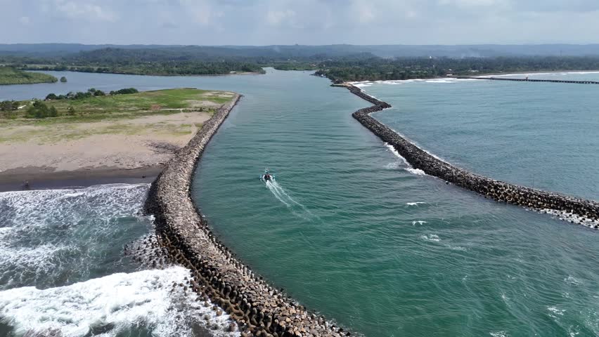 Aerial view of coastal inlet with two boats navigating through a tetrapod breakwater channel surrounded by calm blue-green water and a natural landscape in the background