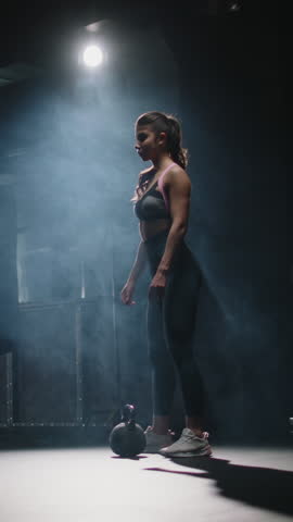 Strong athletic woman lifting kettlebell above head one hand full body view dark gym endurance strength discipline motivation fitness healthy lifestyle vertical slow motion workout