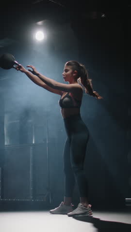 Strong athletic woman lifting kettlebell above head one hand full body view dark gym endurance strength discipline motivation fitness healthy lifestyle vertical slow motion workout