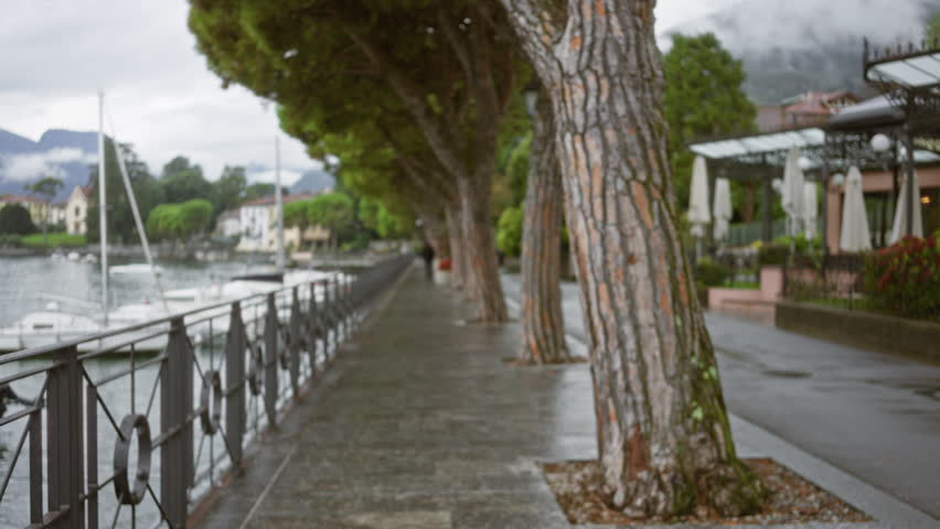 Lake como lakefront promenade in soft bokeh with blurred trees, railing, boats and wet stone walkway street; background backdrop copyspace calm.