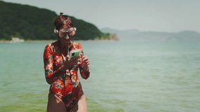 Woman in bright floral swimsuit standing in sea with snorkeling mask and smartphone in waterproof case, smiling while looks at captured photos and videos. Concept digital lifestyle, summer vacation. - Powered by Shutterstock - Get 15% off with code: PIKWIZARD15