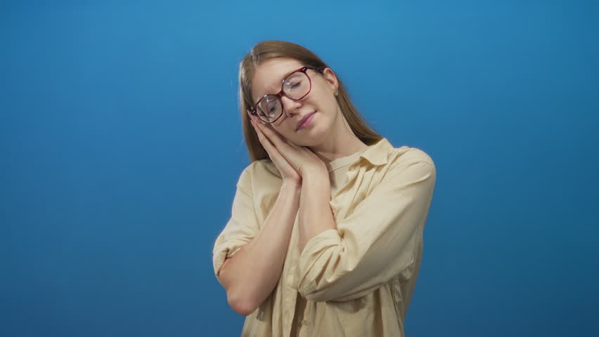 Young woman with glasses and closed eyes resting head on clasped hands in studio; serenity calm repose.