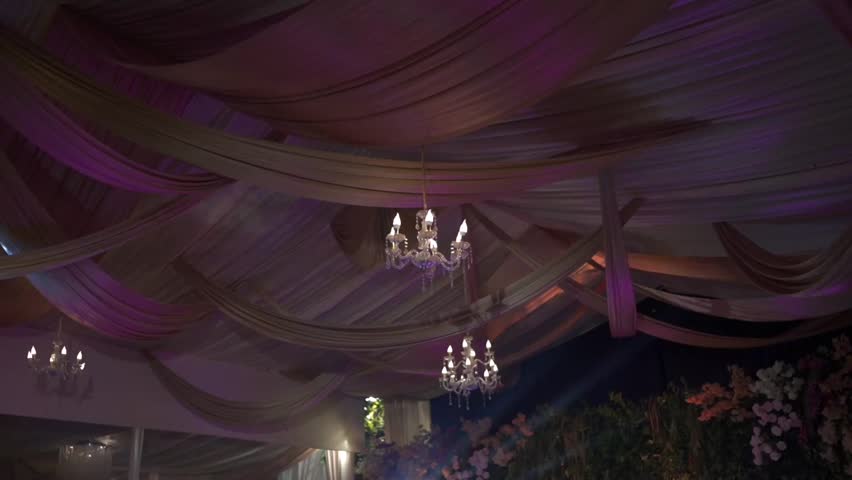 Elegant ceiling decoration with flowing fabric and glowing chandeliers under moving colorful lighting, creating a luxurious event atmosphere.