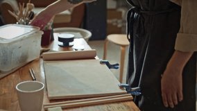 Young female potter in apron kneading raw clay by hand on worktable surrounded by crafting tools and materials in ceramics studio - Powered by Shutterstock - Get 15% off with code: PIKWIZARD15