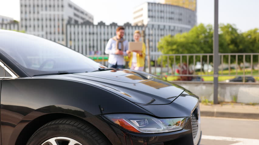 A sleek black car is the focus, with a blurred couple in the background near a city building.