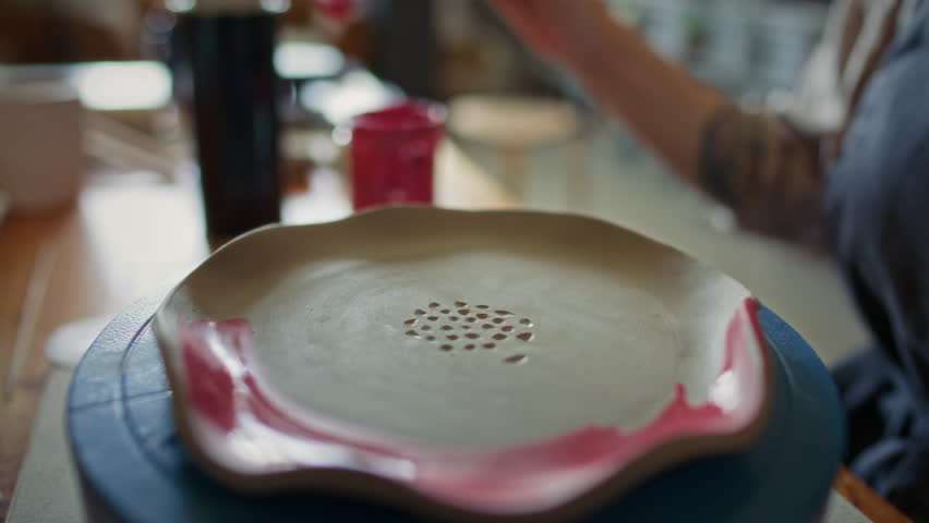 Close-up view of hand of female potter using paintbrush to apply red glaze to edge of handmade ceramic plate with carved pattern placed on turntable in workshop