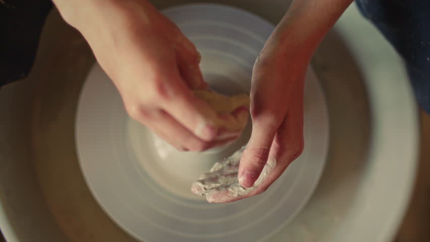 Close-up view of hands of female artisan holding wet clay on spinning pottery wheel during ceramic crafting process. Top down shot