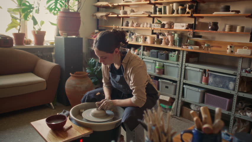 Focused young woman in apron shaping clay on spinning pottery wheel while working in cozy ceramics workshop with various tools and handcrafted items on shelves