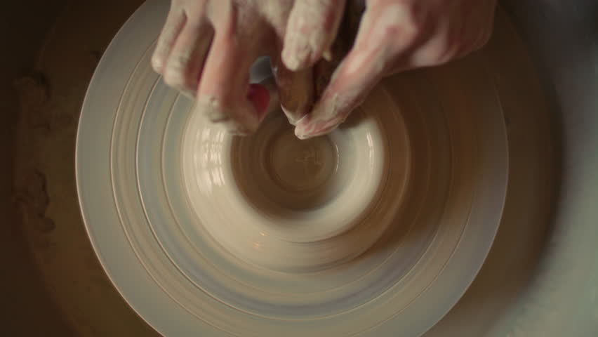 Hands of craftsperson forming clay bowl on spinning pottery wheel in ceramics workshop. Directly above, close-up view
