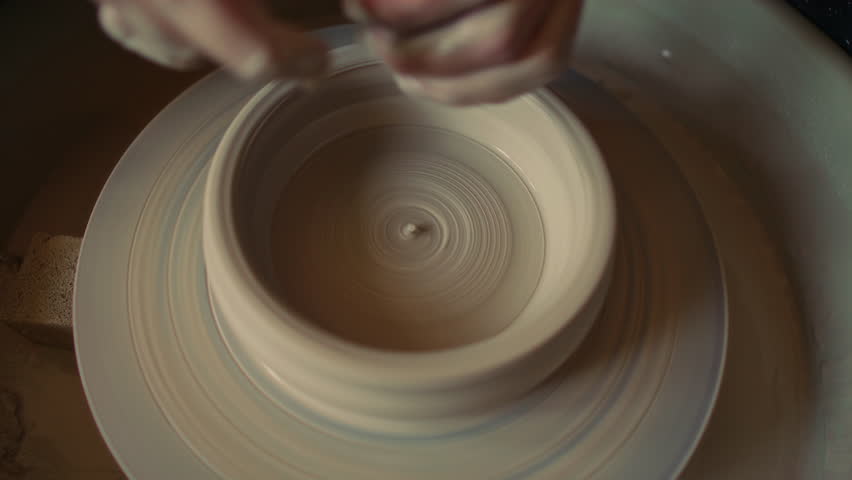Close-up shot of hands refining inner wall of clay bowl with fingers on pottery wheel during ceramic shaping process in workshop