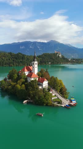 Vertical aerial view of picturesque Lake Bled Island with church and vibrant turquoise water, Bled Castle visible on distant hilltop beneath the Julian Alps in Slovenia