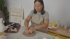 Woman kneading clay with hands at a studio worktable, shaping pottery while wearing an apron and surrounded by carving tools and vases; focused creativity. - Powered by Shutterstock - Get 15% off with code: PIKWIZARD15