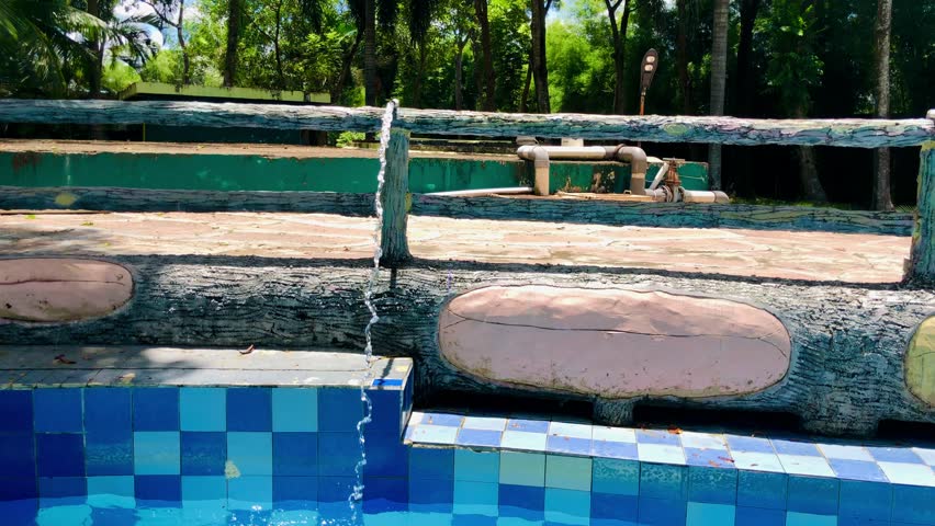 Water flows from a rustic, log-style fountain into a blue-tiled swimming pool in a lush tropical setting, perfect for depicting resort vacation, nature relaxation, or water feature design.