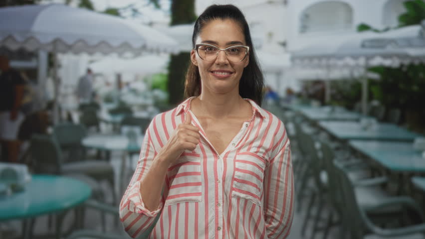 Woman points finger to chest in building terrace restaurant, glasses and striped shirt visible; skepticism doubt.