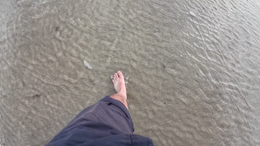 A first-person view of someone walking barefoot through shallow water on Polzeath Beach, UK, as gentle waves ripple over the sand and seaweed drifts nearby, capturing a calm seaside moment.