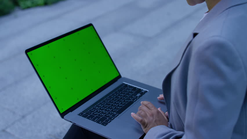 Close-up of a woman using a laptop with a green screen placeholder outdoors, symbolizing online browsing or digital activity.