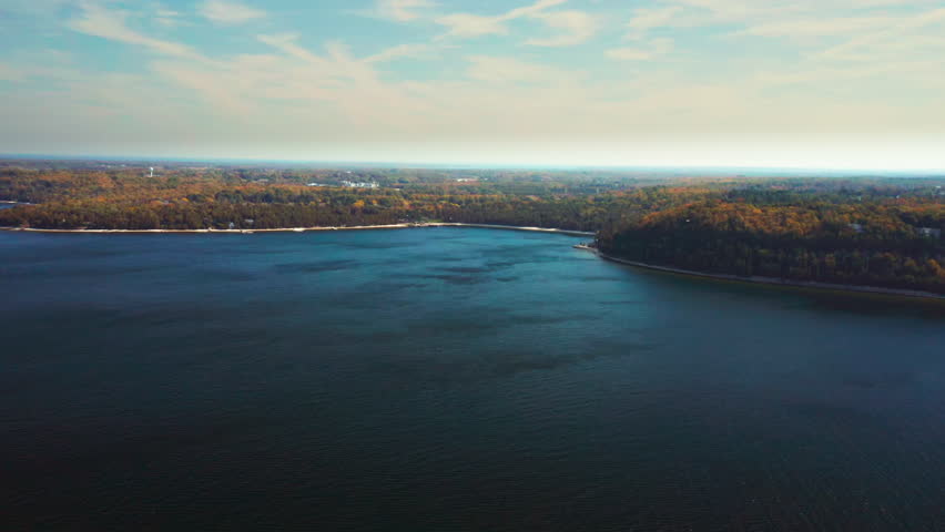 Deep blue waters curve along a forested coastline tinged with autumn hues, stretching toward the horizon beneath streaked clouds and a tranquil afternoon sky.