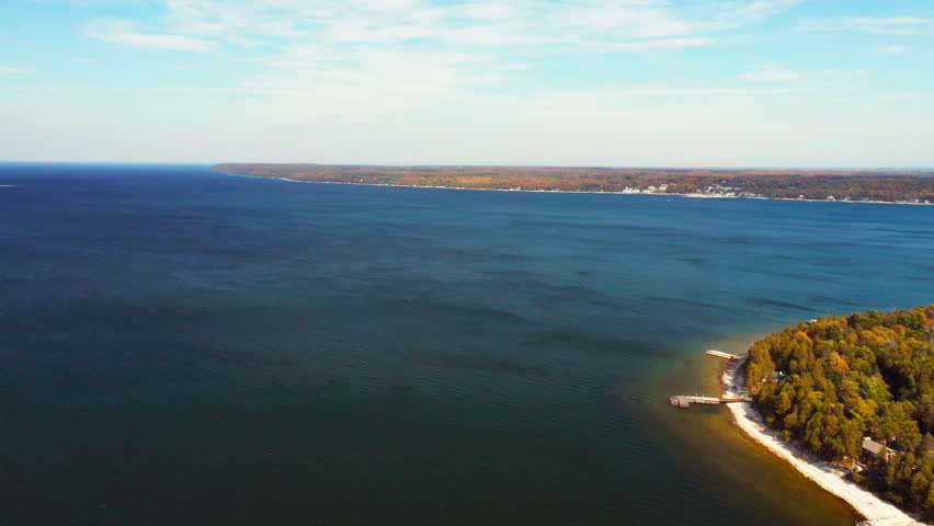 Calm blue water stretches across the frame as autumn-colored trees line a gentle shoreline, where a small wooden pier reaches into the lake beneath bright daylight.
